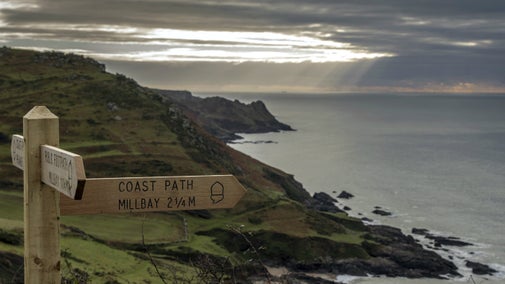 A wooden signpost points to Mill Bay, Devon, with sun breaking through moody, cloudy skies over the sea and dark cliffs in the background